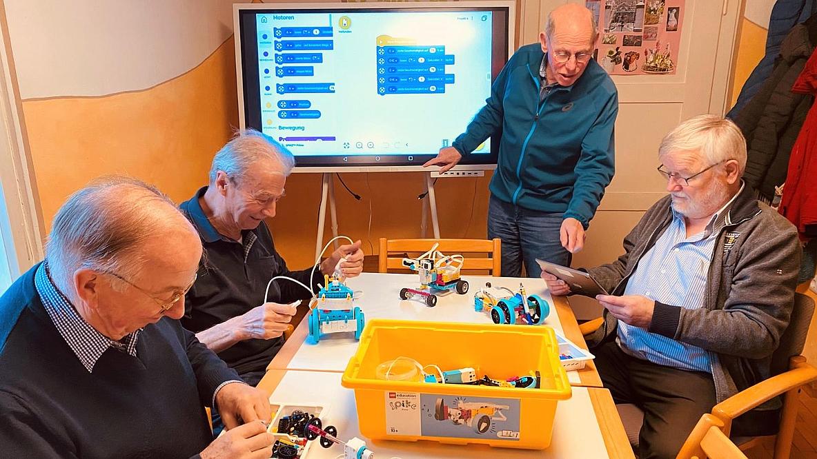Vier ältere Männer bauen Lego-Roboter, im Hintergrund ist ein Bildschirm mit Programmierschritten zu sehen