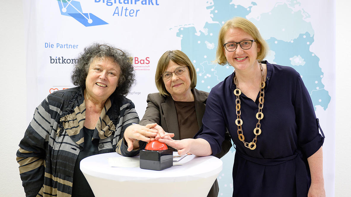 Drei Frauen, Regina Görner, Ursula Nonnemacher und Lisa Paus, drücken gemeinsam auf einen roten Buzzer. Im Hintergrund ist eine Stellwand der Digitalpakt Alter.