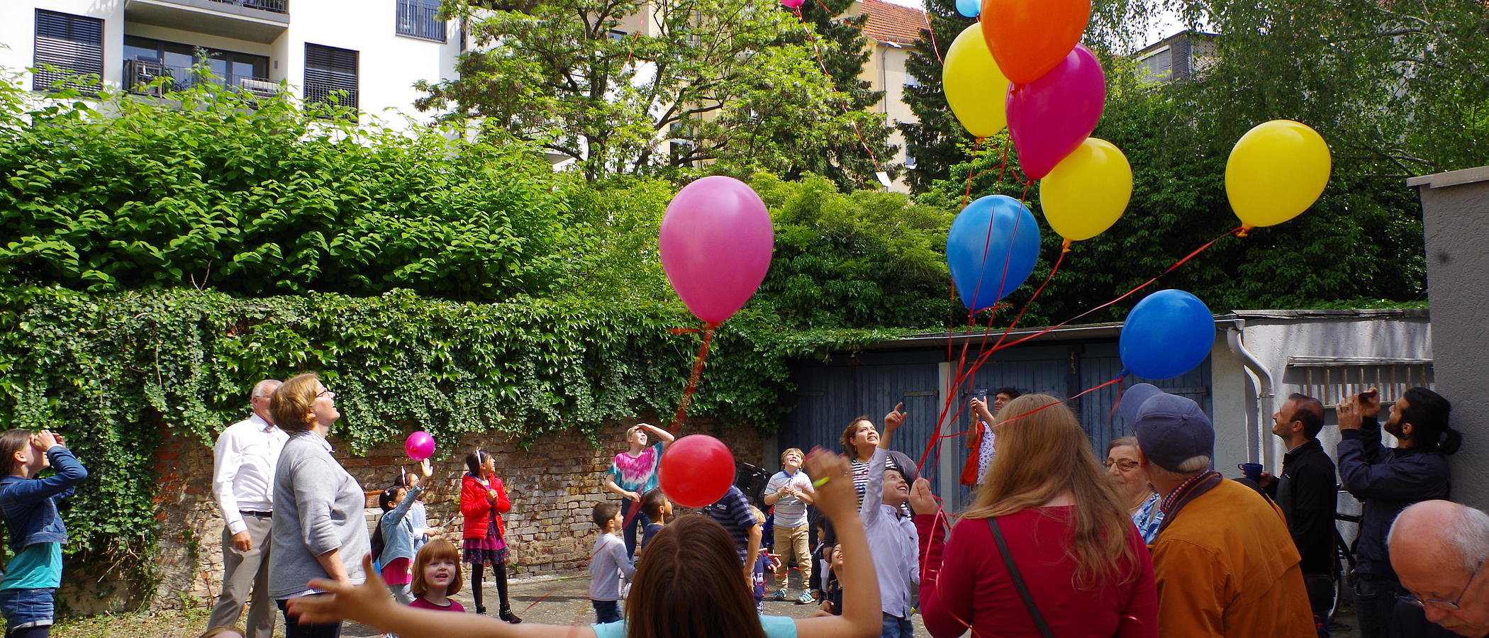 Viele Menschen aller Altersgruppen in einem Hinterhof lassen Luftballons steigen.