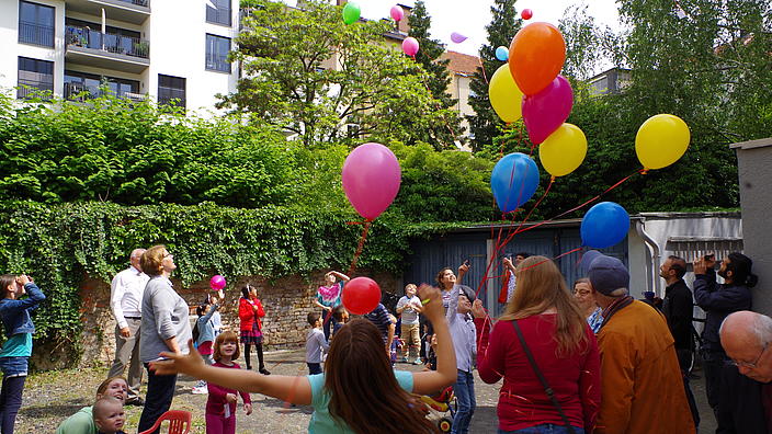 Viele Menschen aller Altersgruppen in einem Hinterhof lassen Luftballons steigen.