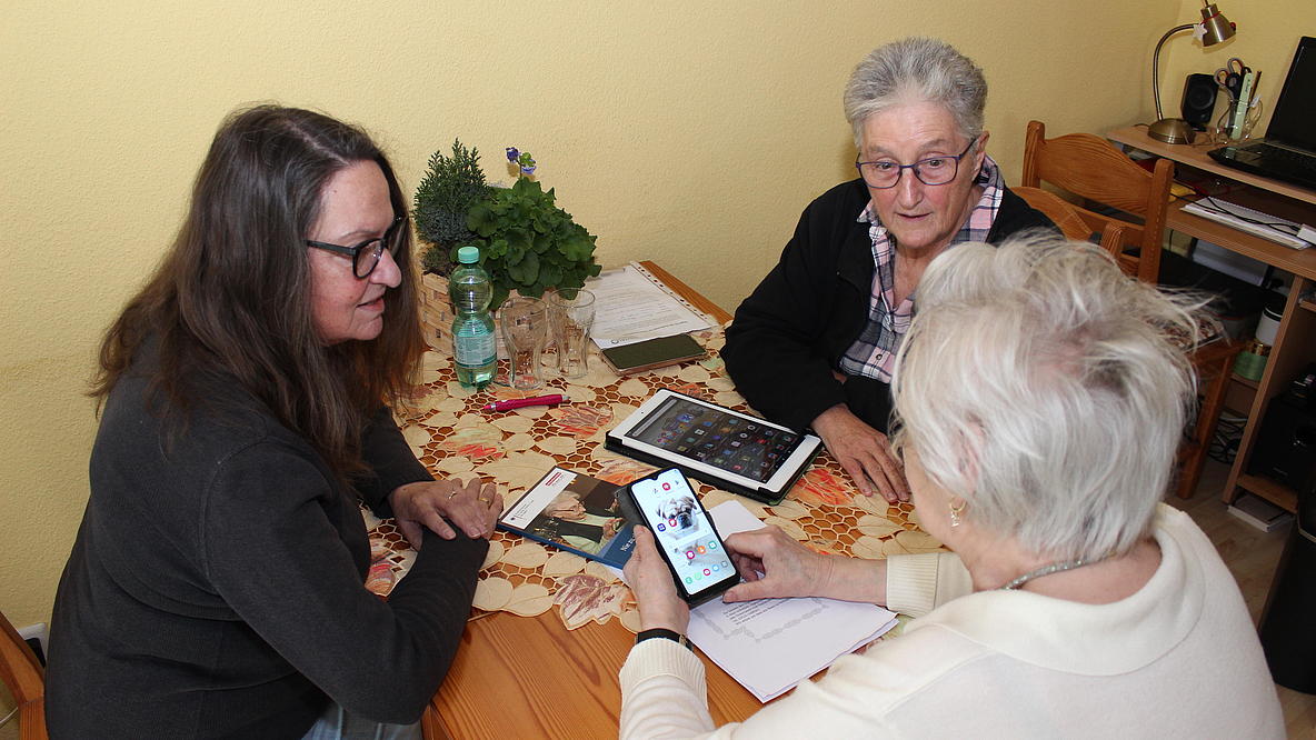 Drei ältere Frauen sitzen am Tisch, eine hält ein Smartphone in der Hand, ein Tablet und eine Broschüre liegen auf dem Tisch. Sie unterhalten sich.