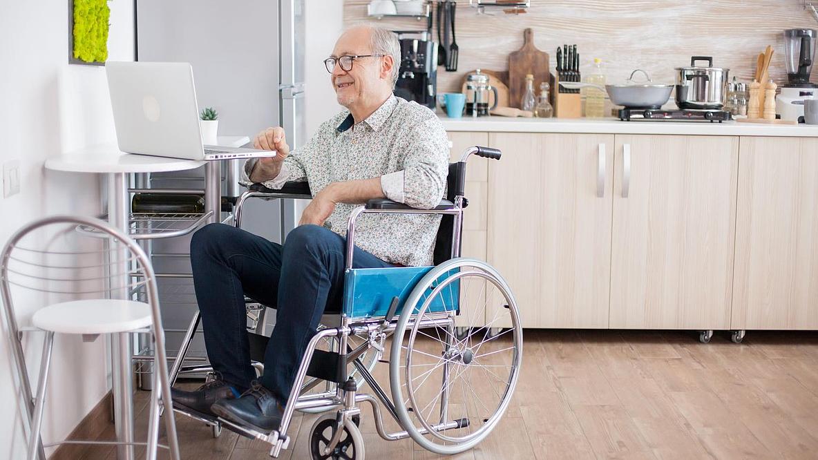 Älterer Mann im Rollstuhl mit Laptop in der Küche.