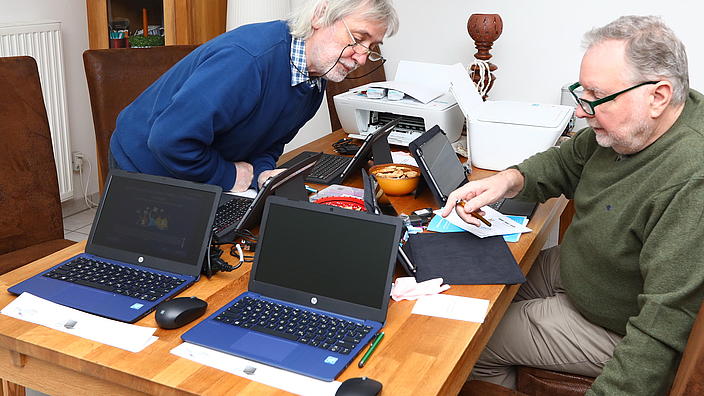 Zwei ältere Herren sitzen mit Laptops am Tisch. Einer beugt sich zu dem anderen Herrn rüber und die besprechen etwas am Laptop.